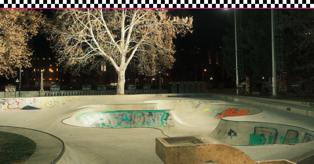 An empty, floodlit concrete skate bowl with graffiti at night under a bare tree, framed by a black-and-white checkerboard stripe and a hot-pink rule